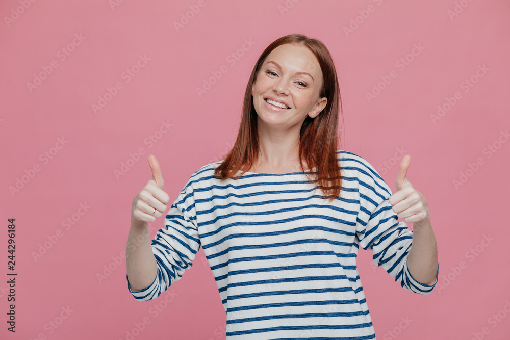 Positive young European woman with reddish hair keeps two thumbs raised, tilts head, dressed in striped clothes, demonstrates agree gesture, wears striped sweater, isolated over pink background Positive young European woman with reddish hair keeps two thumbs raised, tilts head, dressed in striped clothes, demonstrates agree gesture, wears striped sweater, isolated over pink background