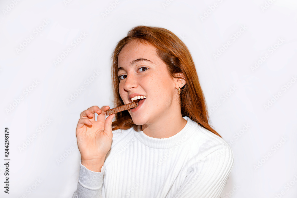 Young red-haired woman, with long hair and freckles, wearing a white shirt on white background. The woman is smiling and eating a chocolate bar. Young red-haired woman, with long hair and freckles, wearing a white shirt on white background. The woman is smiling and eating a chocolate bar.