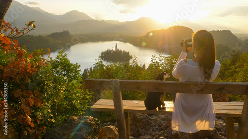 CLOSE UP: Woman taking photos of lake Bled as she sits on bench with her dog.