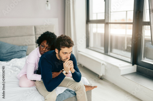 Young displeased caucasian man sitting on bed while his african wife embracing him from the back, trying to comfort. photo