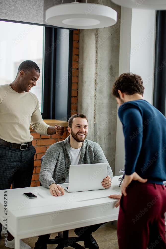 Group of three young multiethnic entrepreneurs standing in the office while debating and discussing new start-up project Group of three young multiethnic entrepreneurs standing in the office while debating and discussing new start-up project