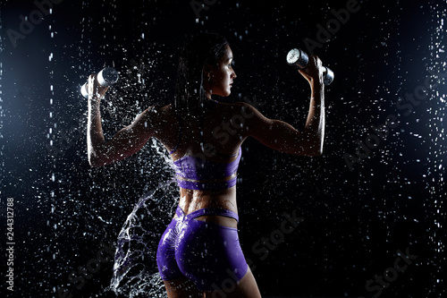 Beautiful young girl in purple sportswear poses with dumbbells in aqua studio. Drops of water spread about her fitness body. The perfect figure on the background of water splashes