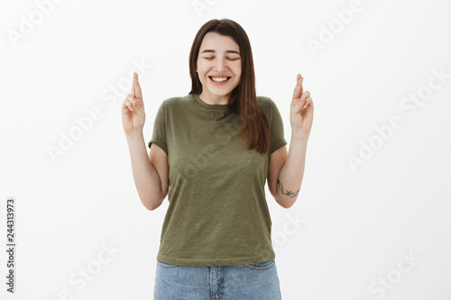 Excited and cute sincere young girl with tattto stooping in thrill and happiness with closed eyes and happy smile, cross fingers for good luck, making wish, praying for dream come true over gray wall photo