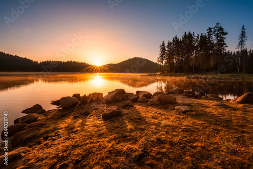 Sunrise on the lake Beautiful sunrise view with sun rays over a mountain lake surrounded by conifers. Shiroka Polyana dam in Rhodopi Mountains, Bulgaria.