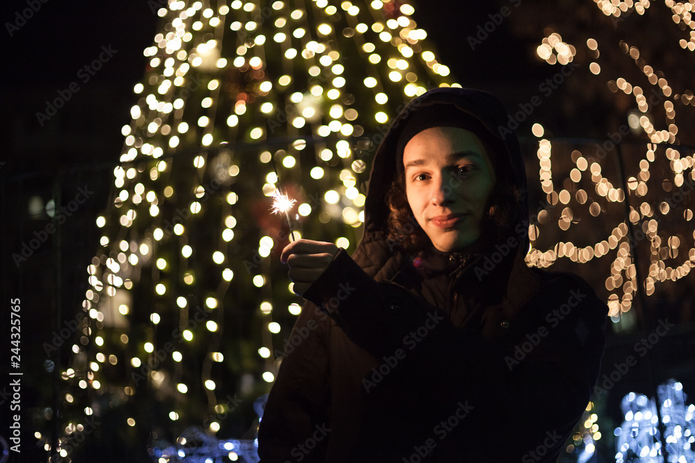 Positive cute boy standing on the street with sparkler in hand. Bright bokeh lights of Christmas tree and garlands on background. Winter holidays mood. Positive cute boy standing on the street with sparkler in hand. Bright bokeh lights of Christmas tree and garlands on background. Winter holidays mood.