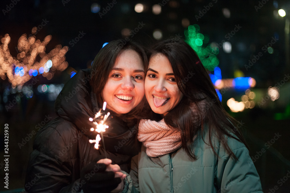 Two happy young girls smiling on the street with sparkler in hand. Bright bokeh lights of Christmas tree and garlands on background. Winter holidays mood.