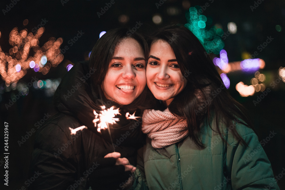 Two happy young girls smiling on the street with sparkler in hand. Bright bokeh lights of Christmas tree and garlands on background. Winter holidays mood.