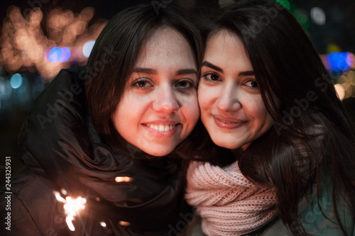 Two happy young girls smiling on the street with sparkler in hand. Bright bokeh lights of Christmas tree and garlands on background. Winter holidays mood. © DubstepWar