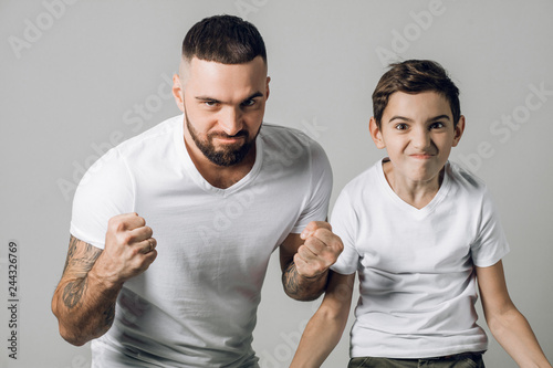 angry frustarated man and his little son in white T-shirts are keeping hands in fists in protective or defensive gesture. protection .isolated on grey background