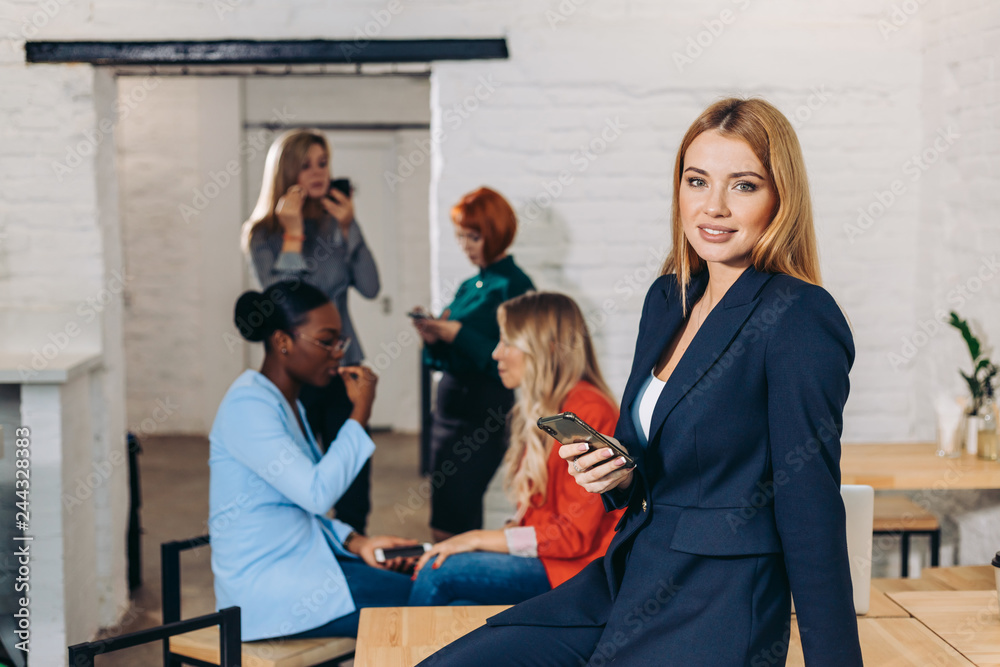 Attractive female manager with long blond hair dressed in blue coat, holding smartphone in hand while sitting in well-lit white office interior with colleagues on background Attractive female manager with long blond hair dressed in blue coat, holding smartphone in hand while sitting in well-lit white office interior with colleagues on background