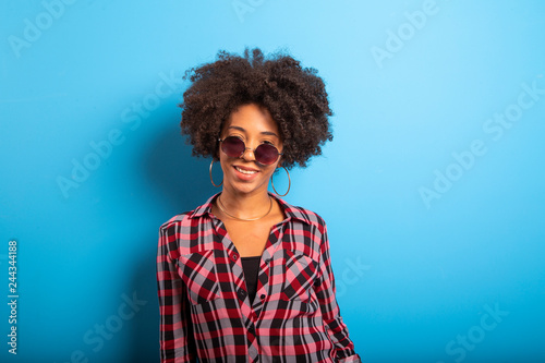 Cheerful attractive afro brazilian girl enjoying life wearing sunglasses On blue wall background - Imagem