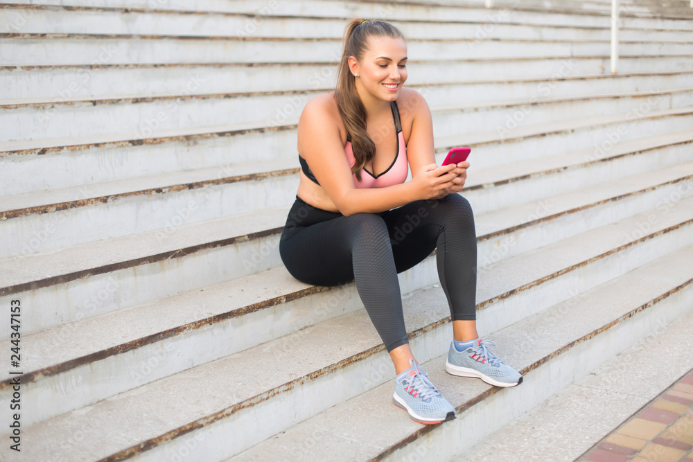 Young cheerful plus size woman in sporty top and leggings sitting on stairs happily using cellphone while spending time outdoor