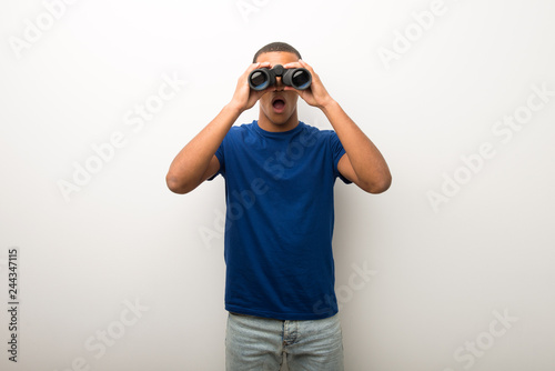 Young african american man on white wall and looking in the distance with binoculars