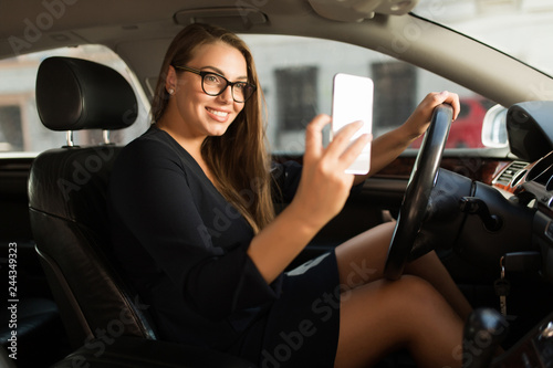Young gorgeous smiling woman in black dress and eyeglasses happily taking photo on cellphone while sitting behind the wheel in car © Anton