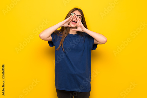 Young woman with glasses over yellow wall shouting and announcing something