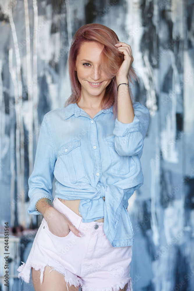 Vertical portrait of pleasant-looking caucasian female with pinkish hairstyle, wearing blue jeans casual shirt and shorts looking happily in camera. People, lifestyle, youth and happiness concept.