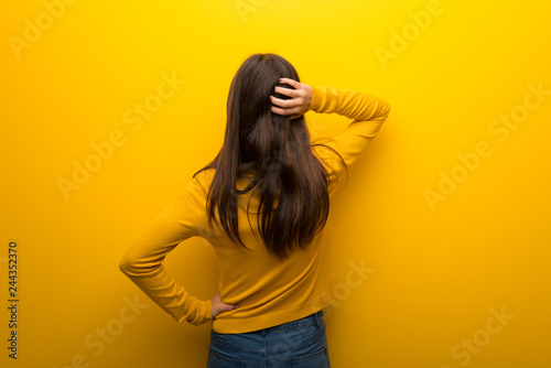 Teenager girl on vibrant yellow background on back position looking back while scratching head photo
