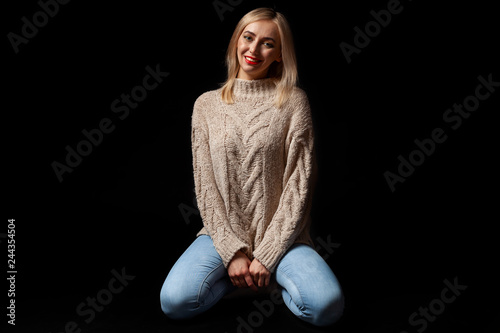 Fototapeta Young beautiful blonde woman in knitted beige sweater, blue jeans,  kneeling on