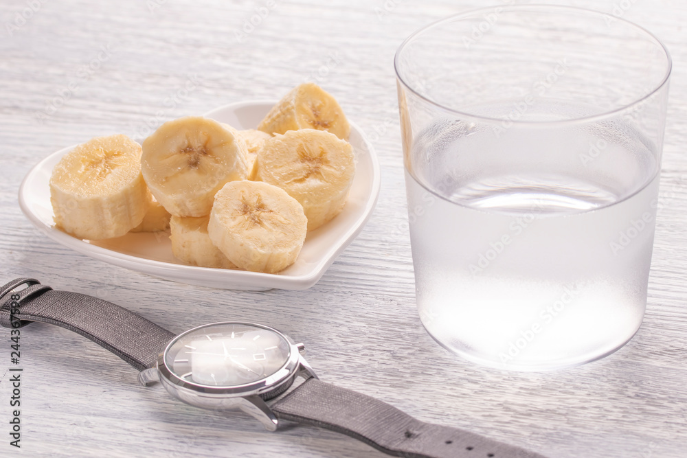 Sliced and whole bananas. On a white plate. On the old wooden table. Misted glass with clear water. Wrist Watch Sliced and whole bananas. On a white plate. On the old wooden table. Misted glass with clear water. Wrist Watch