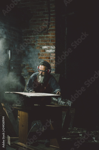 Old bearded carpenter woodworker cutting wood plate with circular saw, process of working, rustic old loft dark style