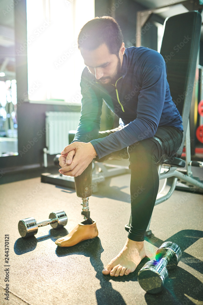 Naklejka premium Sad amputee young bearded man sitting on sort bench and leaning on knees in health club, small dumbbells on floor