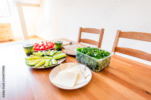 Wooden table setting of healthy vegan vegetarian lunch green vegetables juice, cheese and salad cucumbers tomatoes in home apartment modern house
