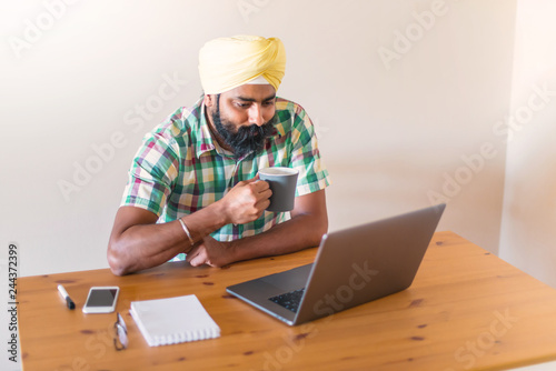 Foto Indian with turban working with his laptop and  holding a cup of coffee