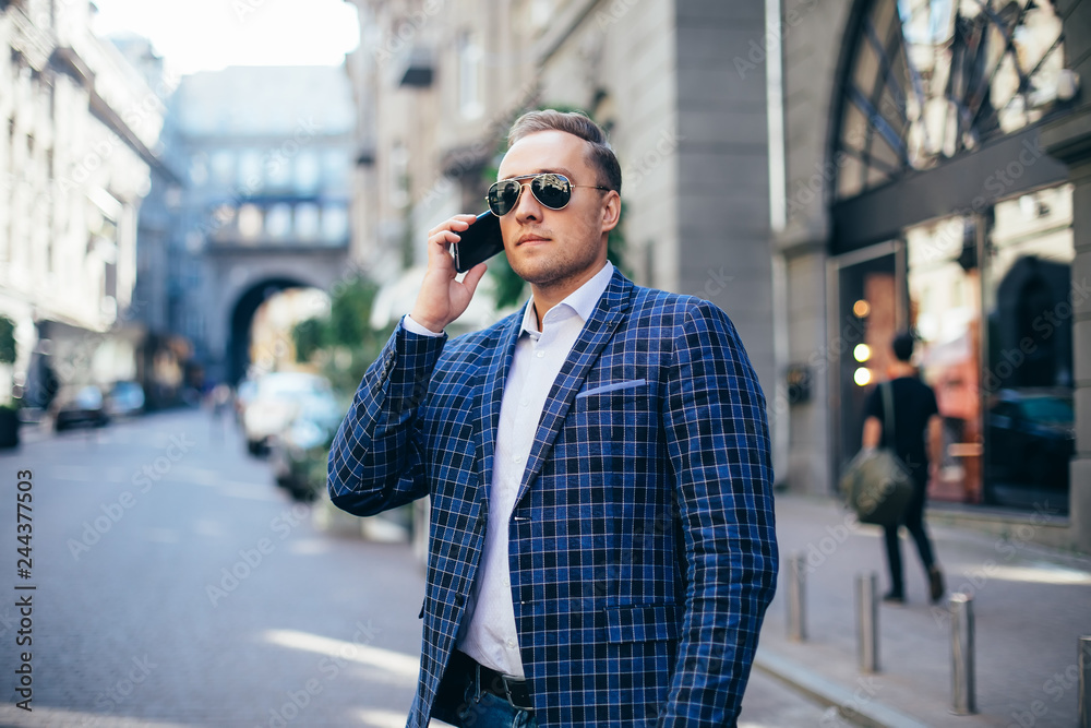 Young stylish man in sunglasses talking on a mobile phone in the city center. wearing a blue jacket and white shirt