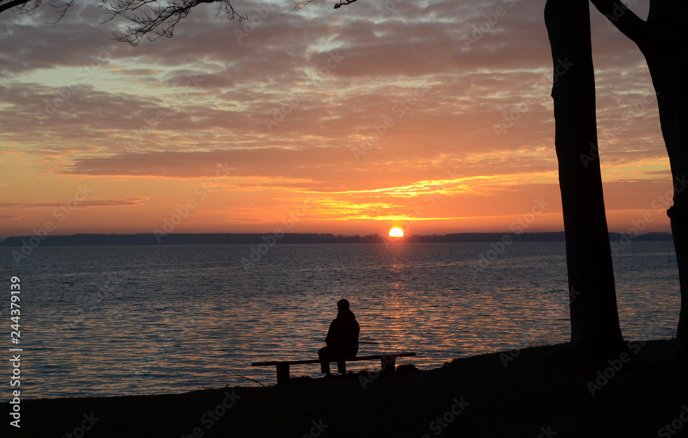 Landscape with silhouette of a person sitting on a bench, sunset and altocumulus clouds over the sea..