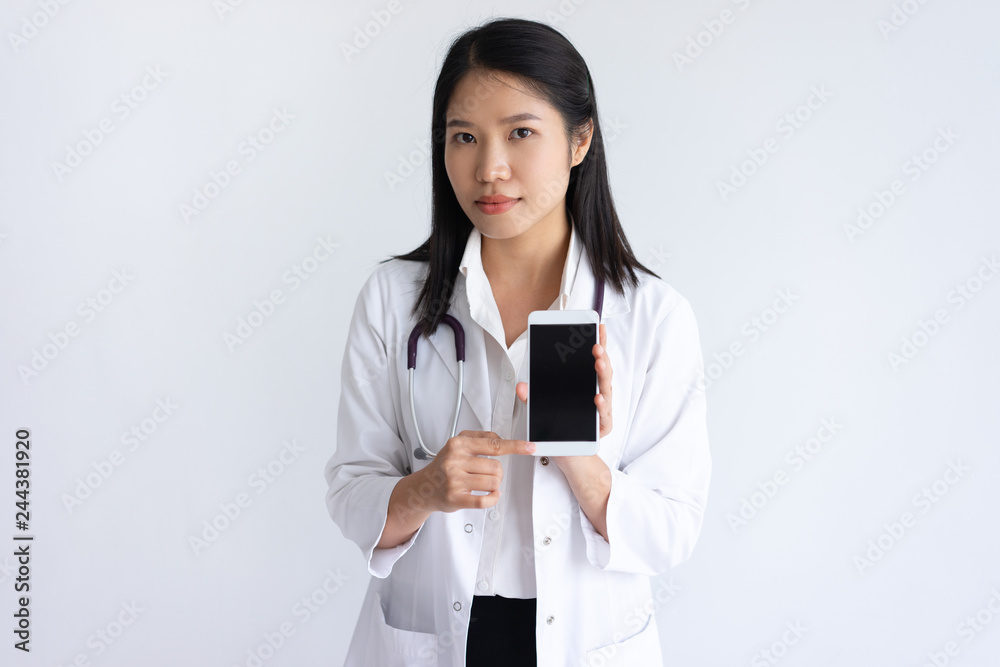 Serious female physician showing smartphone screen. Pretty Asian woman wearing white coat, standing and looking at camera. Healthcare and technology concept. Isolated front view on white background. Serious female physician showing smartphone screen. Pretty Asian woman wearing white coat, standing and looking at camera. Healthcare and technology concept. Isolated front view on white background.