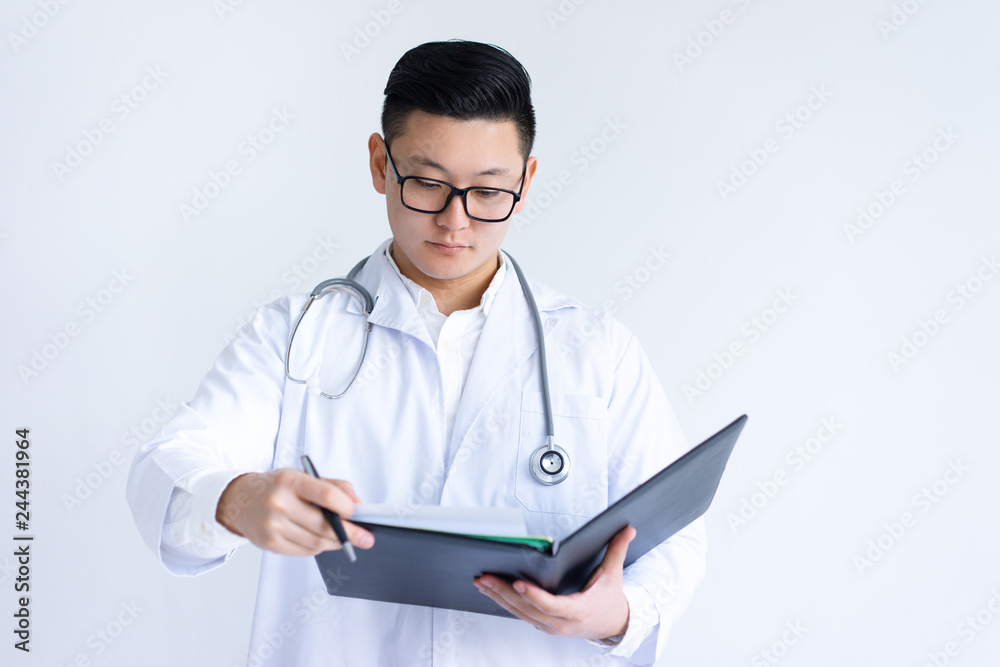 Serious young Asian male doctor working with documents. Man wearing white coat, standing and holding open folder. Doctor occupation concept. Isolated closeup view on white background.