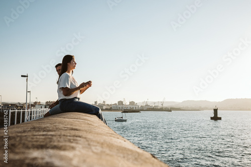 .Young and beautiful couple in love enjoying an afternoon outdoors in Gijón, in Asturias (Spain) overlooking the sea using their mobile phone. Love. Lifestyle.