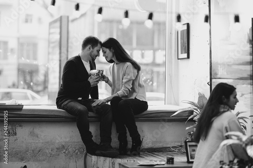 Loving couple dressed in sweaters and jeans is sitting close to each other on the windowsill in a cafe and holding cups in their hands. Black and white photo