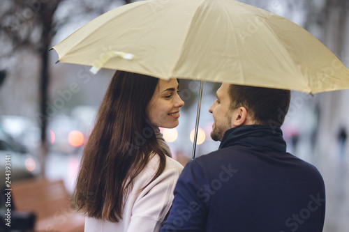 Beautiful couple, guy and his girlfriend dressed in casual clothes stand under the umbrella and look at each other on the street in the rain