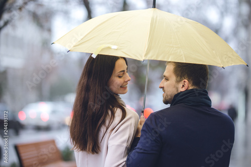 Happy romantic couple, guy and his girlfriend dressed in casual clothes walk under the umbrella and look at each other on the street in the rain.