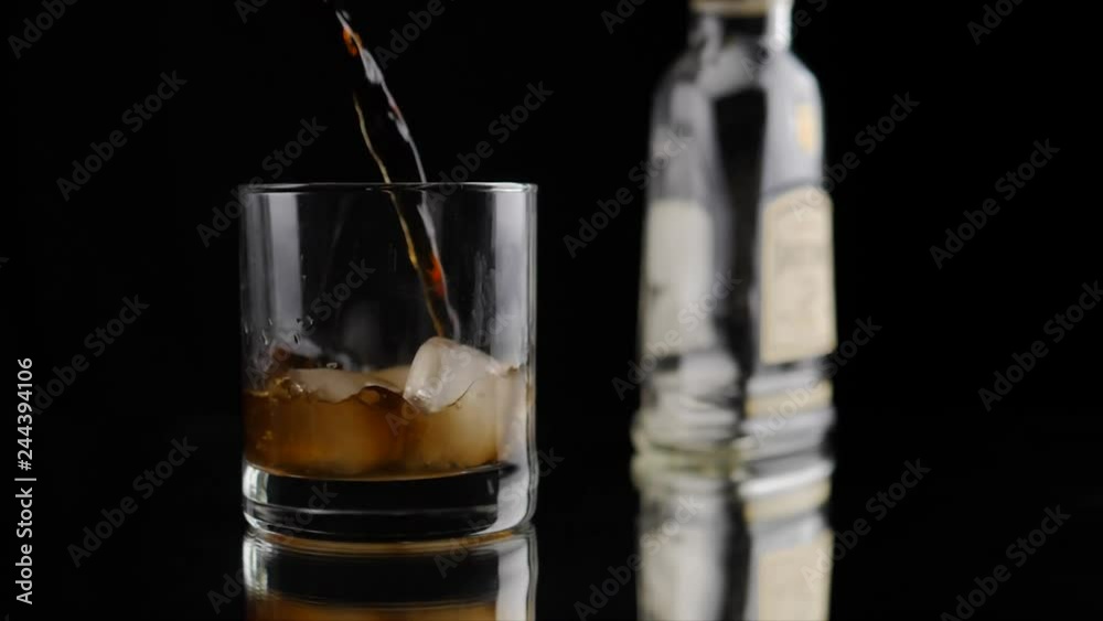Pouring brown color liquid into empty glass with ice. Partly real time, partly slow motion. Shallow depth of field Pouring brown color liquid into empty glass with ice. Partly real time, partly slow motion. Shallow depth of field