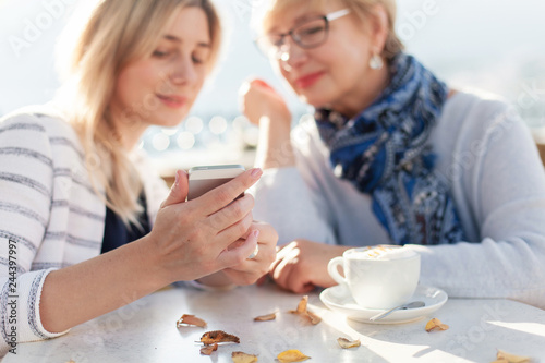 Two beautiful women look at mobile phone and have conversation in cafe with coffee outdoor. Girl shows senior woman a choice on gadget. Blurred background. photo