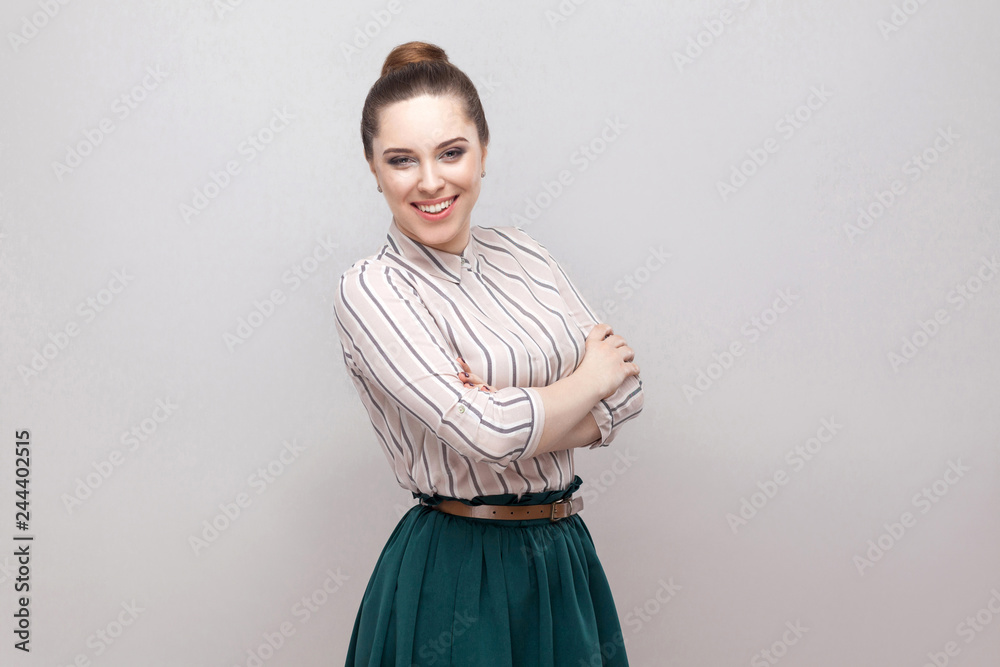 Happy beautiful young woman in striped shirt, green skirt with makeup and ban hairstyle, standing with crossed hands and looking at camera with toothy smile. studio shot, isolated on grey background. Happy beautiful young woman in striped shirt, green skirt with makeup and ban hairstyle, standing with crossed hands and looking at camera with toothy smile. studio shot, isolated on grey background.