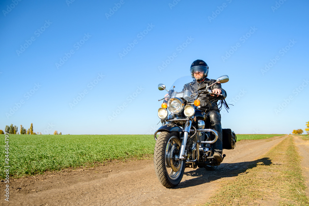 Motorcycle Driver Riding Custom Chopper Bike on Autumn Dirt Road in the Green Field. Adventure Concept. Motorcycle Driver Riding Custom Chopper Bike on Autumn Dirt Road in the Green Field. Adventure Concept.