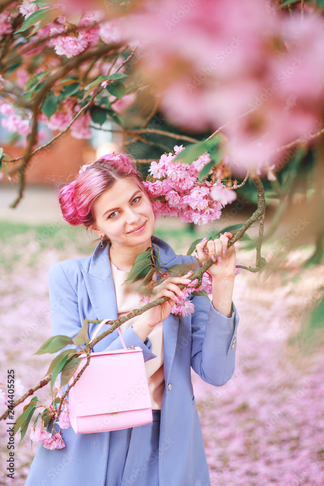 Fototapeta premium Beautiful girl with pink hair in a fashionable blue pantsuit standing next to colorful flowers.