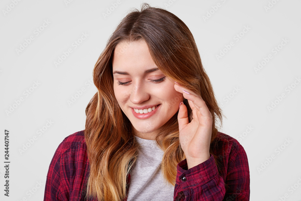 Headshot of cheerful woman with toothy smile, keeps gaze down, has wavy dyed hair, holds hand near cheek, wears checkered shirt, has tender look, isolated over white background. Joy concept. Headshot of cheerful woman with toothy smile, keeps gaze down, has wavy dyed hair, holds hand near cheek, wears checkered shirt, has tender look, isolated over white background. Joy concept.