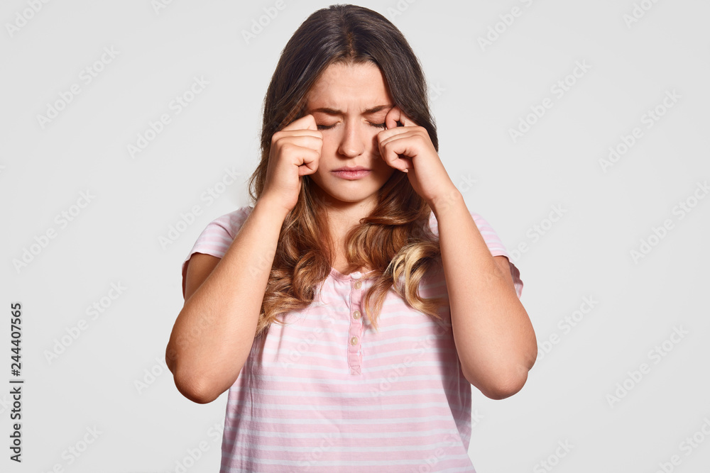 Photo of sleepy European woman rubs eyes, wears casual domestic clothes, wants to have rest, has long dark hair, isolated over white background. People, tiredness, sleeping and rest concept. Photo of sleepy European woman rubs eyes, wears casual domestic clothes, wants to have rest, has long dark hair, isolated over white background. People, tiredness, sleeping and rest concept.
