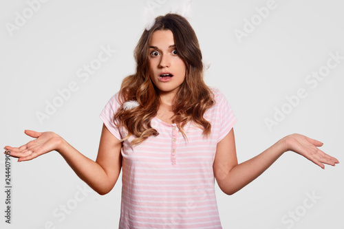 Hesiatnt beautiful woman spreads hands, wears pyjamas, has feathers on head, feels doubt, shocked to oversleep work, doesnt know what to do, isolated over white background. So what to do now? © sementsova321
