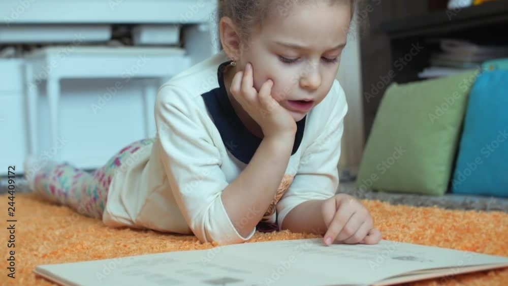 Cute Little Girl with Curly Hair, in children's pajamas, lying on the floor on a knitted carpet and herself reading a children's book. Little girl reading a book hugging her soft baby doll. Cute Little Girl with Curly Hair, in children's pajamas, lying on the floor on a knitted carpet and herself reading a children's book. Little girl reading a book hugging her soft baby doll.