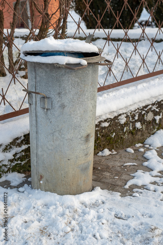 Trash bin at the entrance gate. Metal container covered with snow.