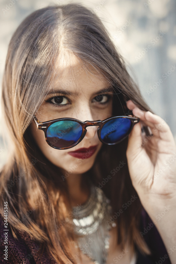 Stylish hipster girl smiling in sunny street on background of wooden wall. Portrait of boho girl in cool outfit and sunglasses posing in sunlight and shadow. Summer vacation and travel Stylish hipster girl smiling in sunny street on background of wooden wall. Portrait of boho girl in cool outfit and sunglasses posing in sunlight and shadow. Summer vacation and travel