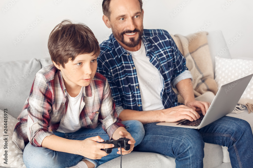 Father and little son at home sitting on sofa dad typing on laptop looking curious at boy playing game console excited