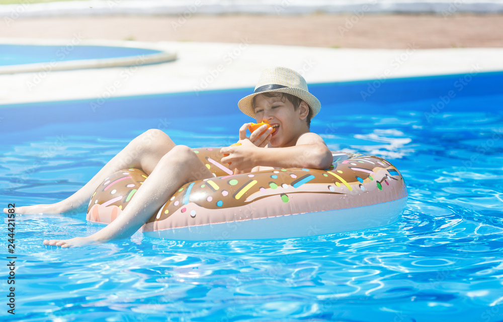 Cute child boy on funny inflatable donut float ring in swimming pool with oranges. Teenager learning to swim, have fun in outdoor pool at resort. Water toys for kids. Healthy sport activity, vacation 