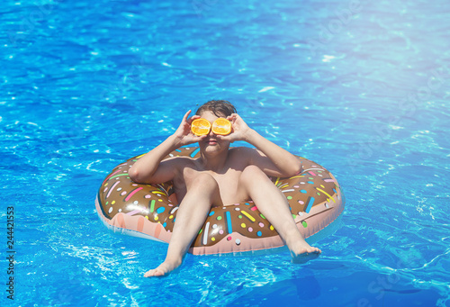 Cute child boy on funny inflatable donut float ring in swimming pool with oranges. Teenager learning to swim, have fun in outdoor pool at resort. Water toys for kids. Healthy sport activity, vacation 