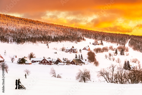 Wonderful view of the calm, snowy and landscape, hillside with beautiful mountain cottages, alleys, road and forest, silhouette of people with a stroller and golden cloudy sky in the background
